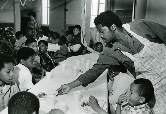 Charles Bursey hands plate of food to a child seated at Free Breakfast Program.