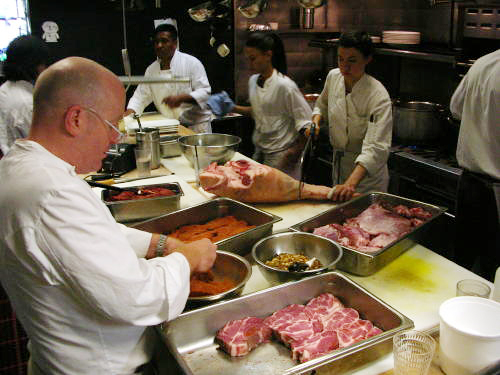Oliveto Chef Paul Canales seasons pork cutlets while Sous Chef Kelsey Bergstrom cuts into a pork leg with a hand saw.