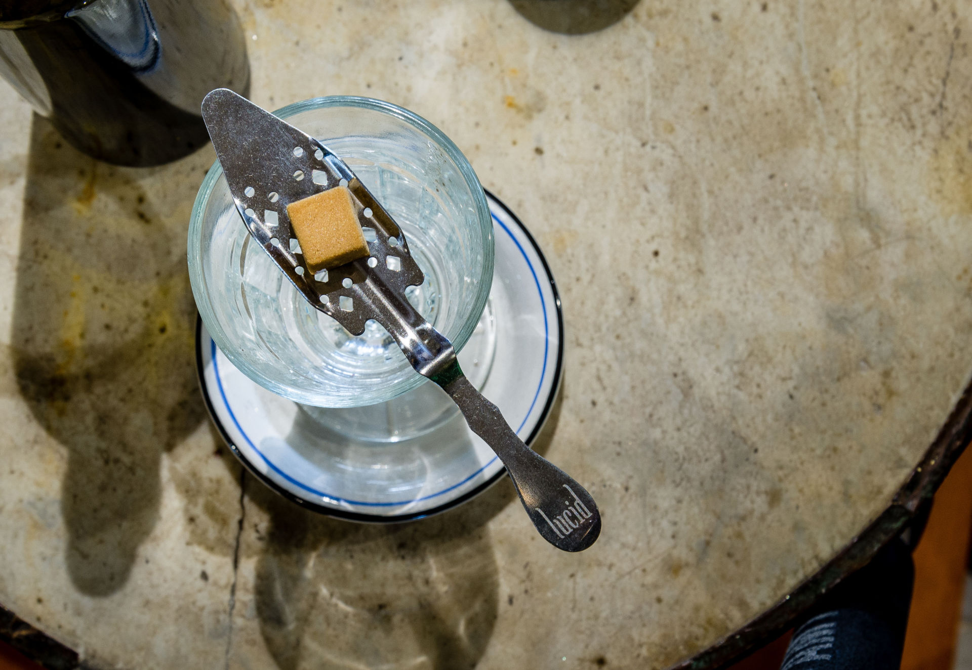 A sugar cube is cradled by a slotted spoon balanced on top of a glass of absinthe.