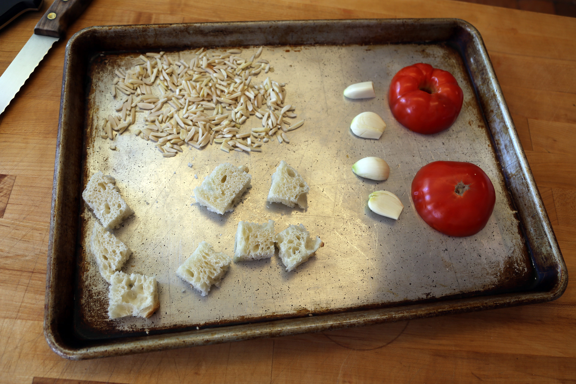Place the tomato, cut side down on one side of a rimmed baking sheet and arrange the garlic next to it. On the other side, add the bread and almonds.
