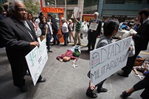 As part of a national day of action against border militarization, faith leaders, workers and community groups held a protest and dramatazation of migrant deaths, outside of the office of one of the top six companies that currently profits from U.S Border contracts. In this photo protesters march around three people pretending to lie dead, representing those that they say have died at the hands of the U.S. Customs and Border Control. Other names of people that they say have died were read out loud. (Deborah Svoboda/KQED)
