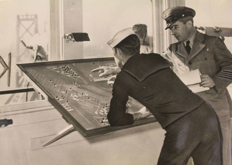 Navy signal corpsman F.R. English at the berth board receiving instruction from Chief Signalman Stanley Jones. The operations of the signal tower were military secrets until Dec 27th, 1945. One of the towers of the Bay Bridge's western span is visible in the background.
