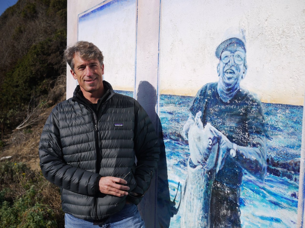 Mike Wallace used to bring his high school surf team to practice at Martins Beach. Here, he stands next to a mural depicting the beach's former owners. (Amy Standen/KQED)