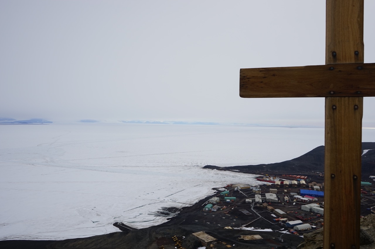 View of McMurdo Station from Observation Hill, where a cross has been erected to the Robert Falcon Scott expedition, whose members died on their return from the South Pole. Polar Star is docked. Note the turning basin at left and the channel, dimly visible, which runs out to open water in the distance.