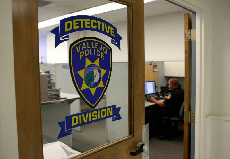 Viewed through a glass wall with blue-painted badge insignia and logo for "Detective Division" of officer sitting at desk in front of computer.