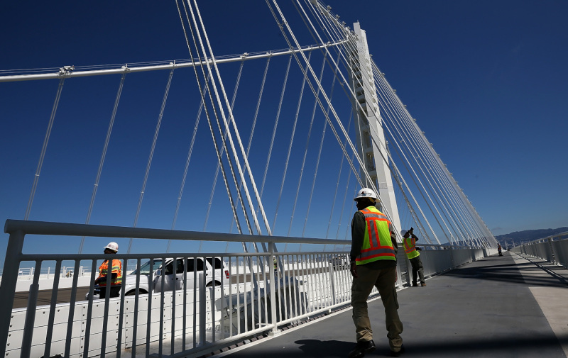 A worker in a flourescent yellow safety vest stands on the Eastern Bay Bridge span bike path, with a towering white spire and support cables in front of him.