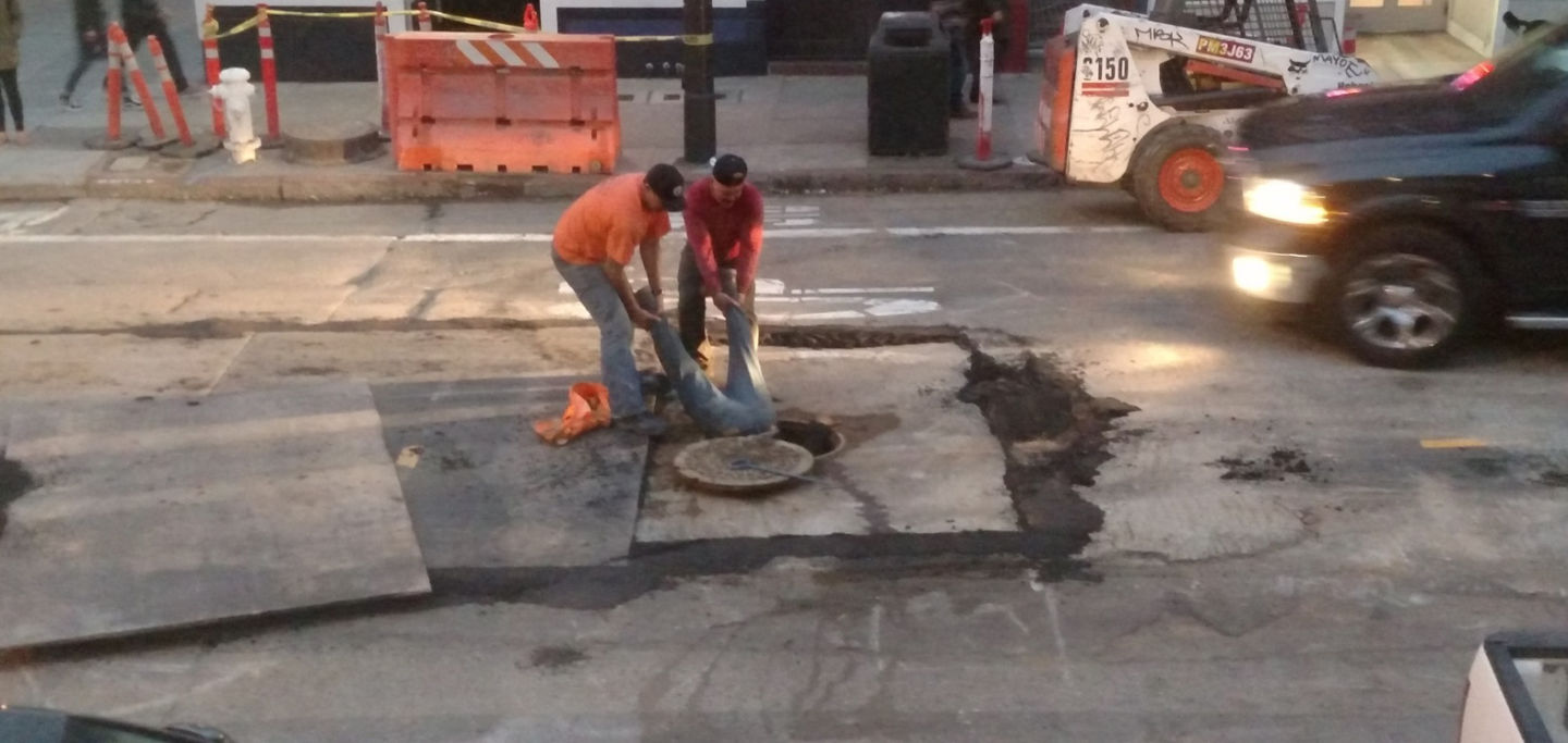 Two workers from Synergy Project Management hold a foreman by his ankles as he works in a Haight Street manhole on July 21, 2015.  San Francisco Public Works