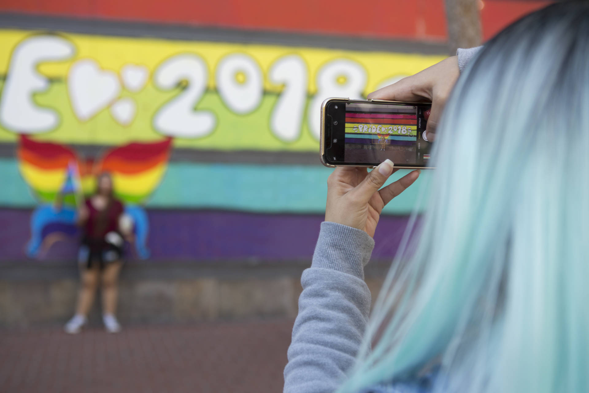 Keila Cisneros takes a photo of her sister, Marina, in front of a mural on Market street that says "Pride 2018." "I'm here to support my sister who just came out as bisexual and my best friend who is totally gay," said Cisneros with a giggle.  Anne Wernikoff/KQED