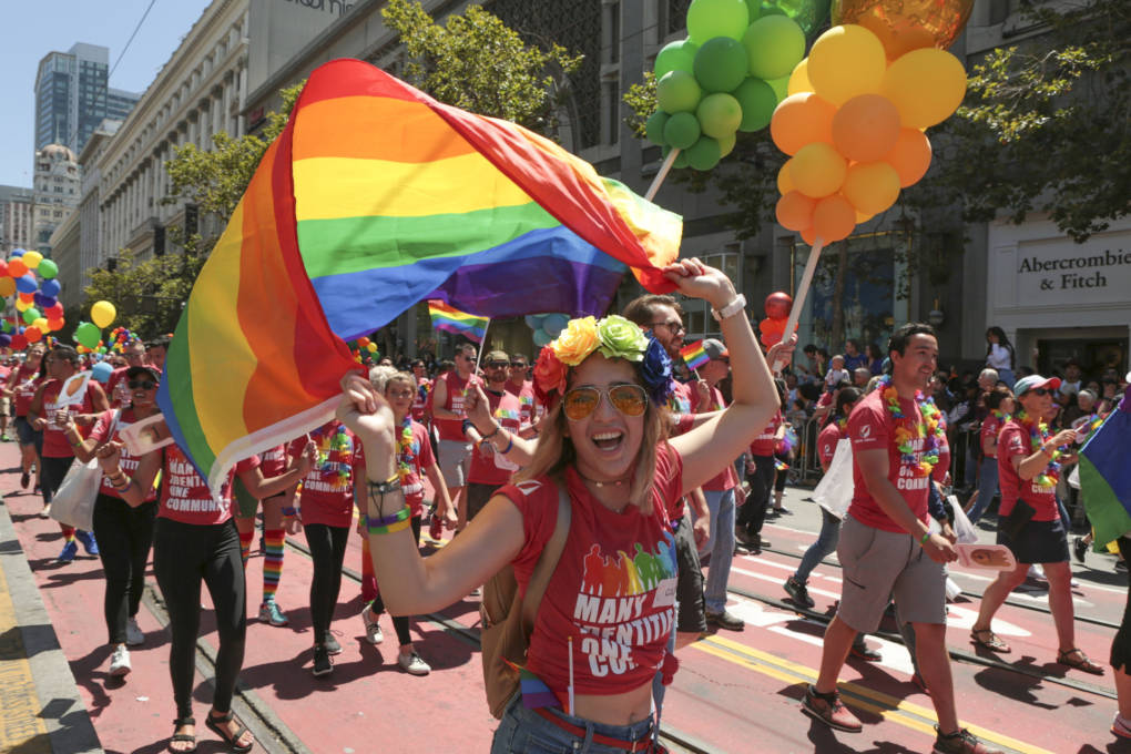 A scene of the Pride parade on Market Street in San Francisco. In the foreground is a white woman wearing rose-colored glasses and holding a rainbow flag with both hands above her head. She is surrounded by other marchers and towers of green and orange balloons. It is sunny, and this is a joyful scene, as the woman is smiling broadly.