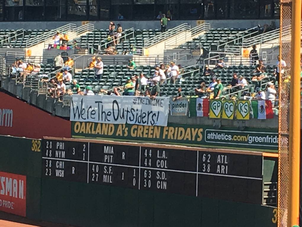 A sign that reads "We're the Outsiders!" hangs from the bleachers of a sports stadium with people in the stands.