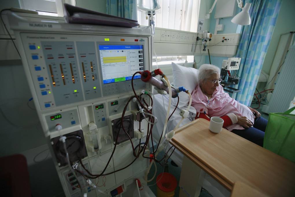 A hospital room, where in the foreground a large, square machine with a screen and lights and tubes coming out of it connect to a woman sitting beyond it, with white hair and glasses, a pink button-up shirt and blue pants.
