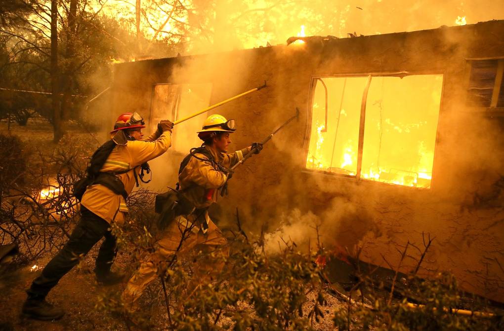 Two people dressed in firefighter gear holds equipment poles to a burning structure.