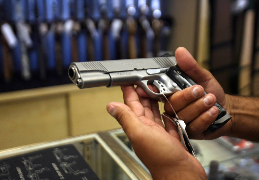 A person holds a handgun with both hands with other firearms in the background.