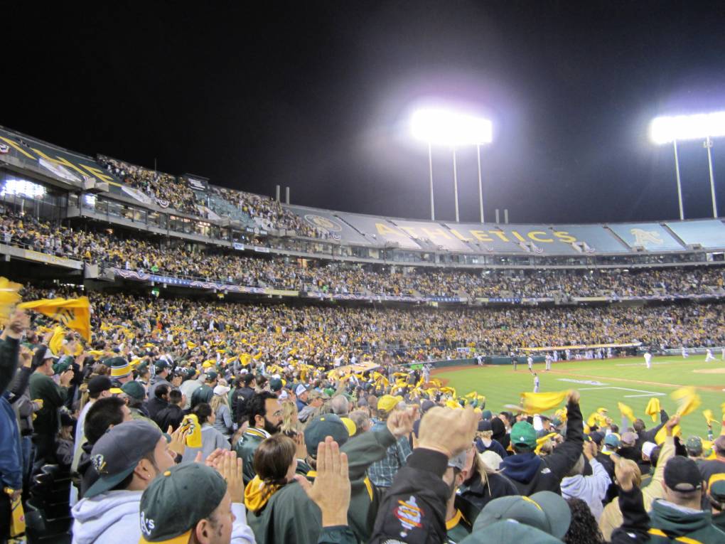 Many people clapping at an A's baseball game with bight lights and dark sky in the distance