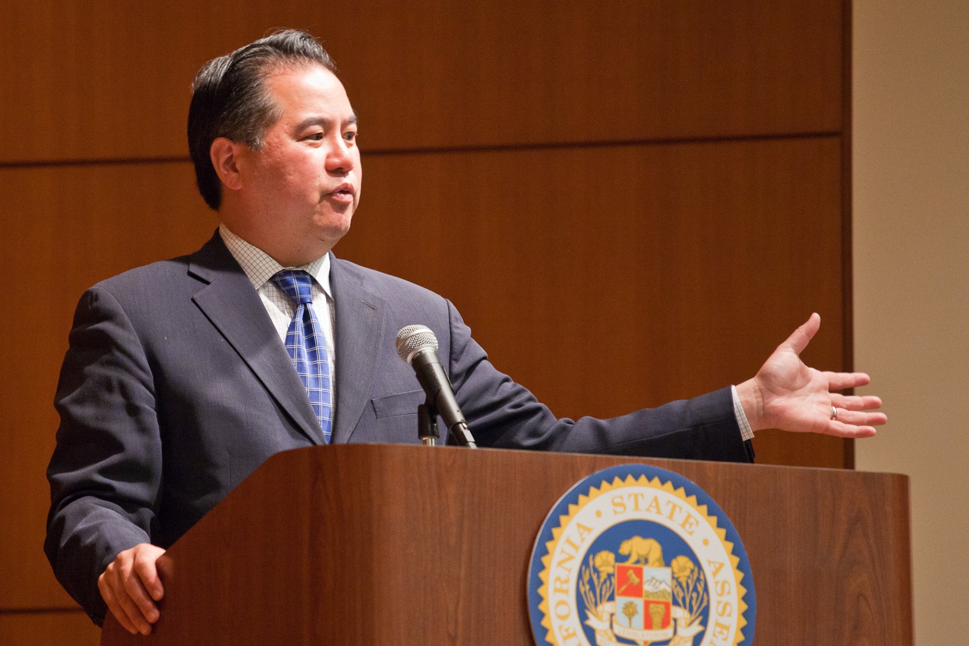 An Asian man in a suit and tie speaks from behind a dais with a California emblem.