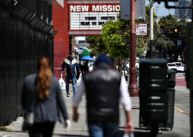 Two people photographed from behind walking down what is recognizably Mission Street in San Francisco, because of the New Mission theater sign in the background, in focus.