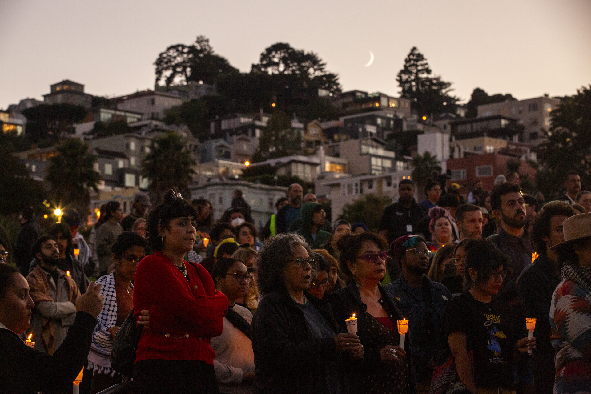 A large crowd stands in silence in a large park, and looks away from the camera. Many are holding candles. All have serious expressions.