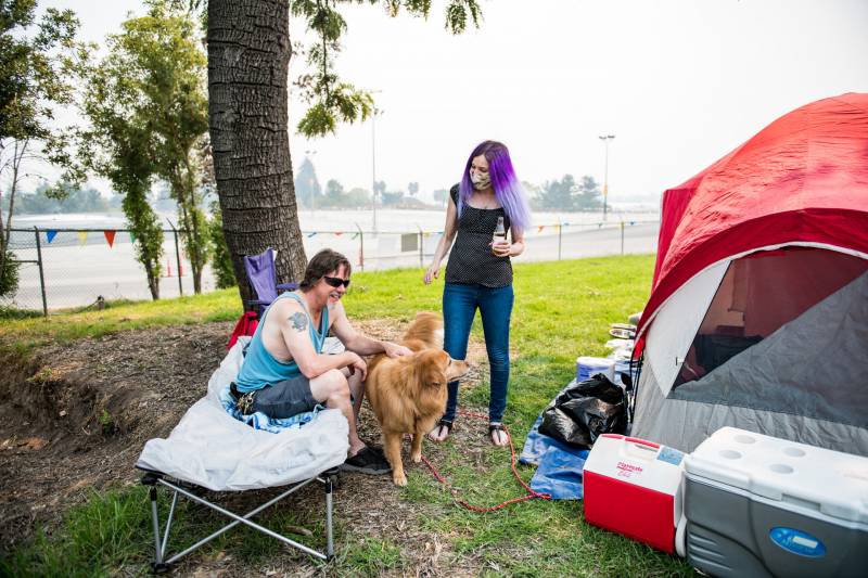 Man sits on a cot petting a dog, beside a standing woman with purple hair and a red tent on a grassy field