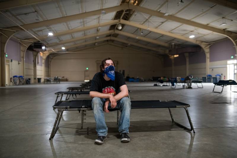 Man with blue bandana over lower half of face rests on cot in hangar-like building