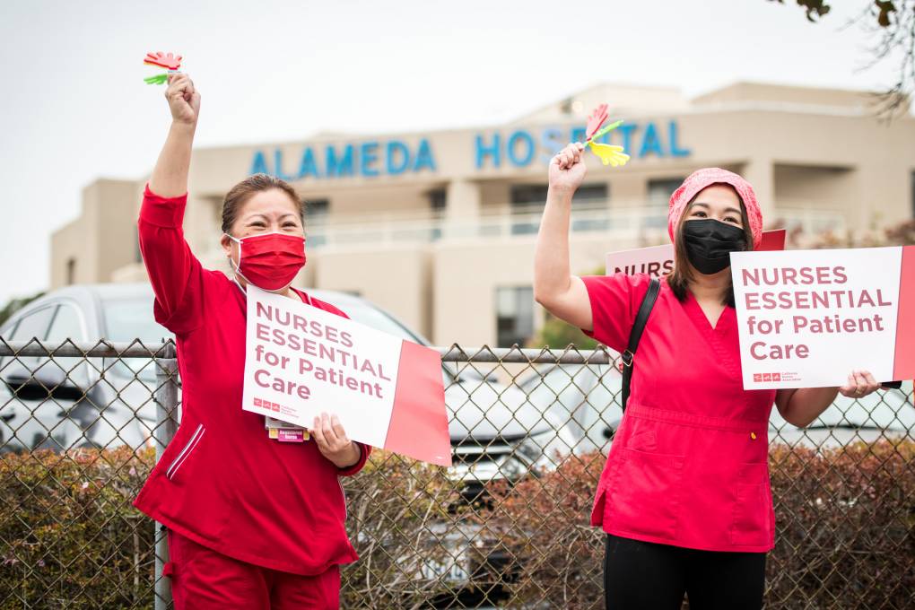 Two nurses wearing red wave and hold signs that say, "Nurses Essential for Patient Care."