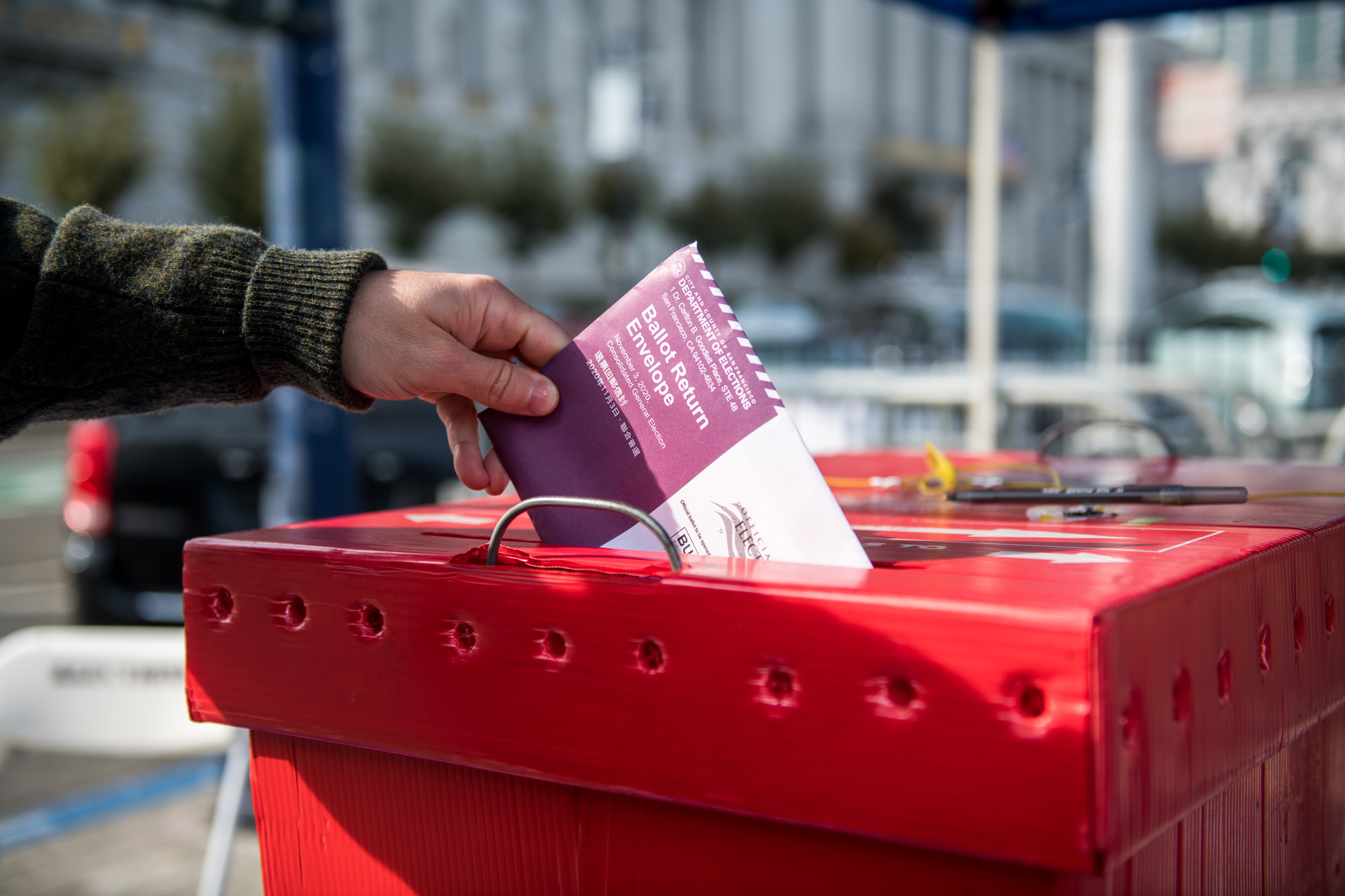 Hand holding ballot drops it in red cardboard ballot box