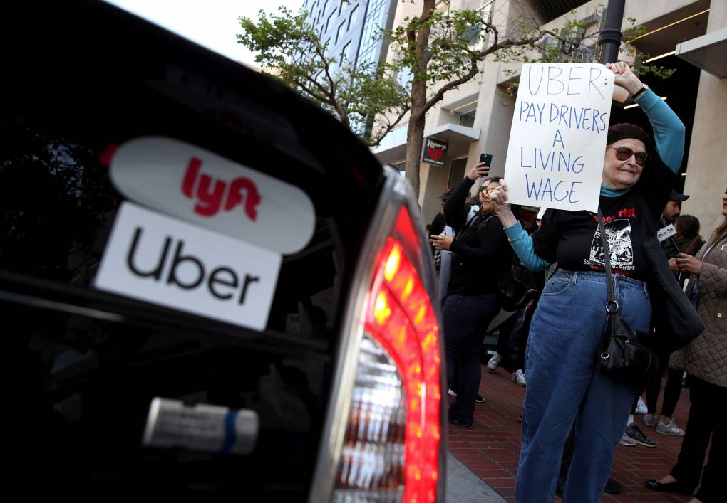 Protesters stand on a sidewalk in front of an office, and next to a car with Lyft and Uber stickers on the back. A woman holds a sign that says: Uber: 'Pay Drivers a Living Wage.'