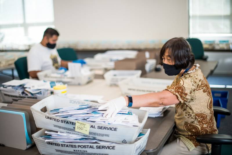 A woman in short sleeves and a mask sits at a fold-out table in front of baskets of mail.