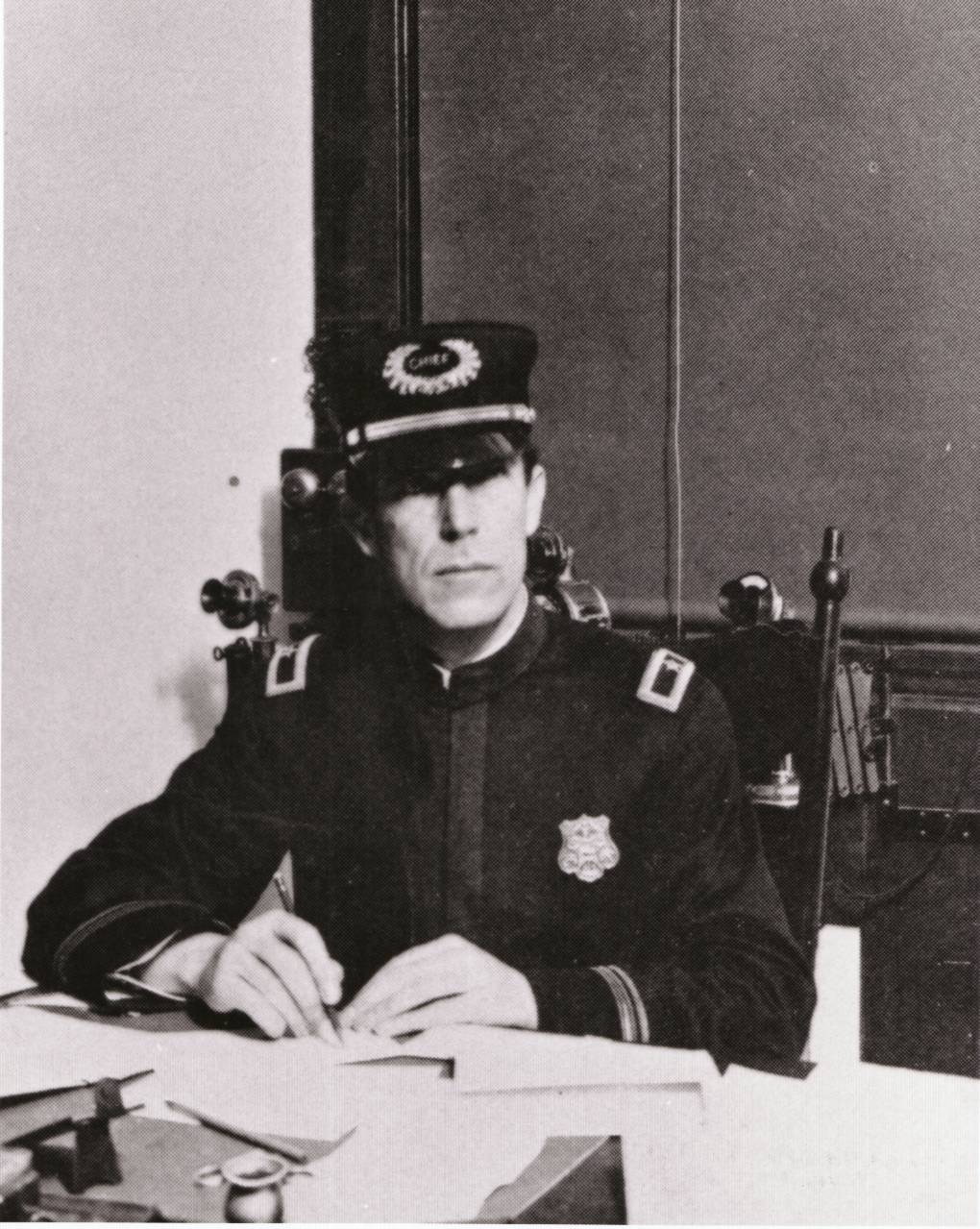 Portrait of August Vollmer sitting at his desk wearing a dark uniform and his chief's hat.