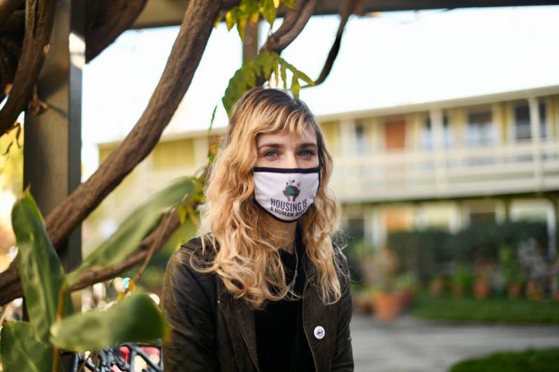 A woman standing in front of apartments, wearing a mask that reads 'housing is a human right'