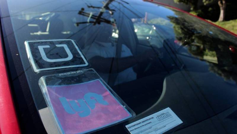 A view of the dashboard of a car with Uber and Lyft logos on the window. Inside sits a man in the driver's seat.