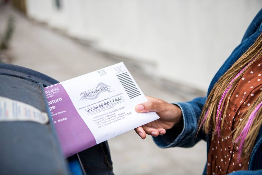 A hand slides a ballot into a blue postal box.