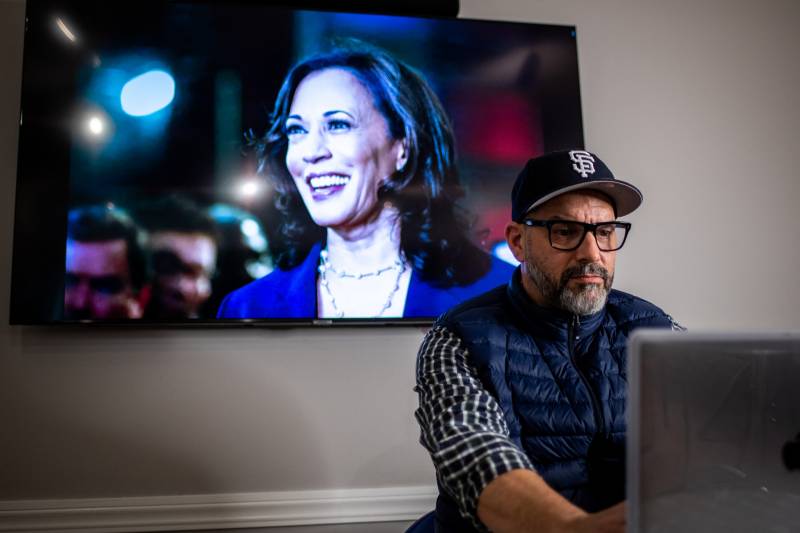 Billy Lemon works on a computer with Vice President-elect Kamala Harris on a television screen in the background.