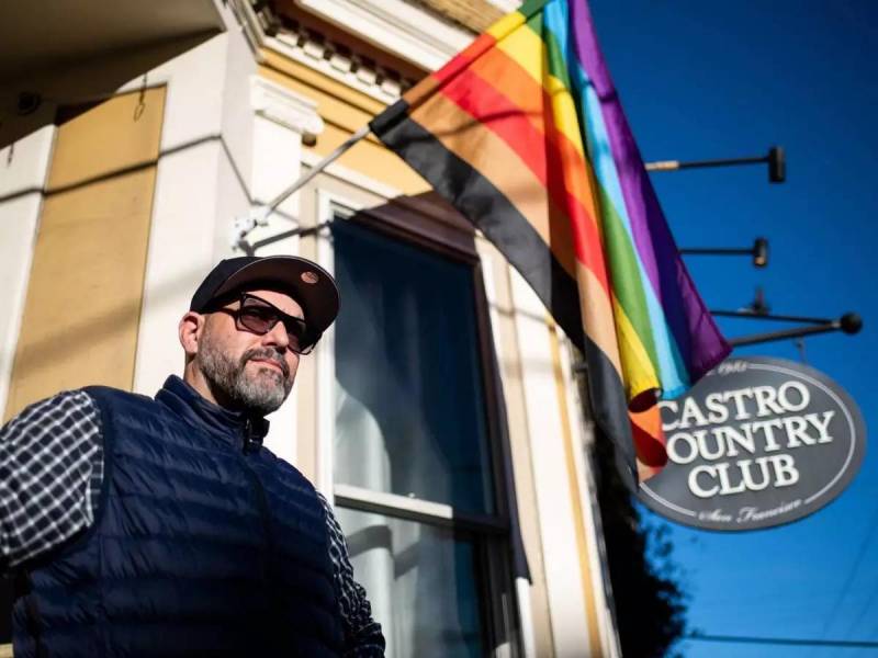 A man in sunglasses and a hat stands beneath a Pride flag outside a Victorian with a sign that says Castro Country Club.