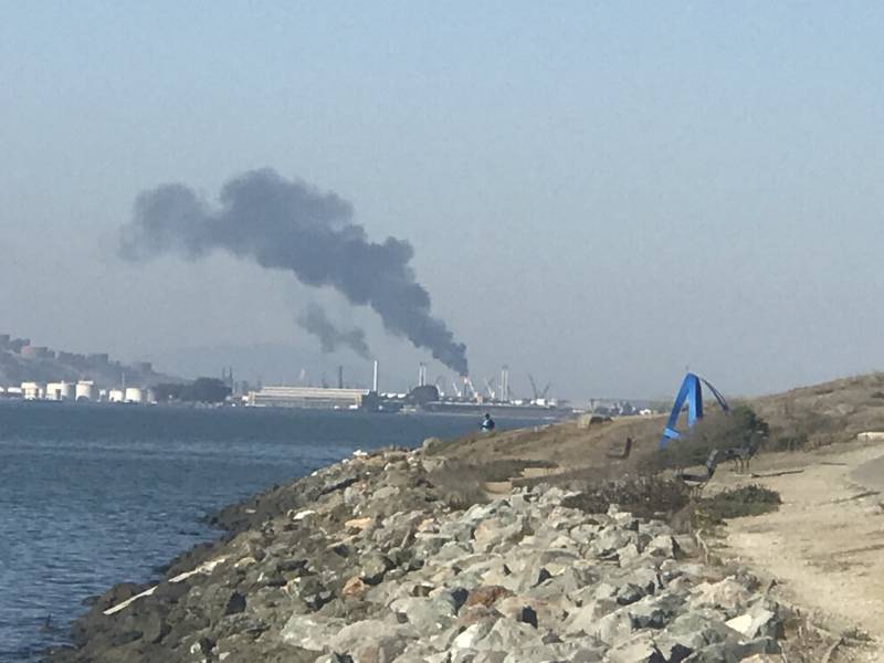 View from a rocky shoreline across the bay to a refinery spewing a massive plume of smoke.