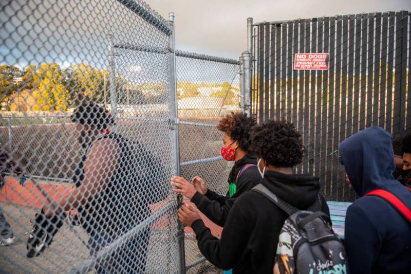 Students wearing masks and backpacks walk through a chain-link fence.
