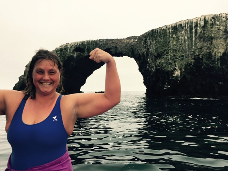 Amy Gubser flexes her arms with ocean and coastline behind her