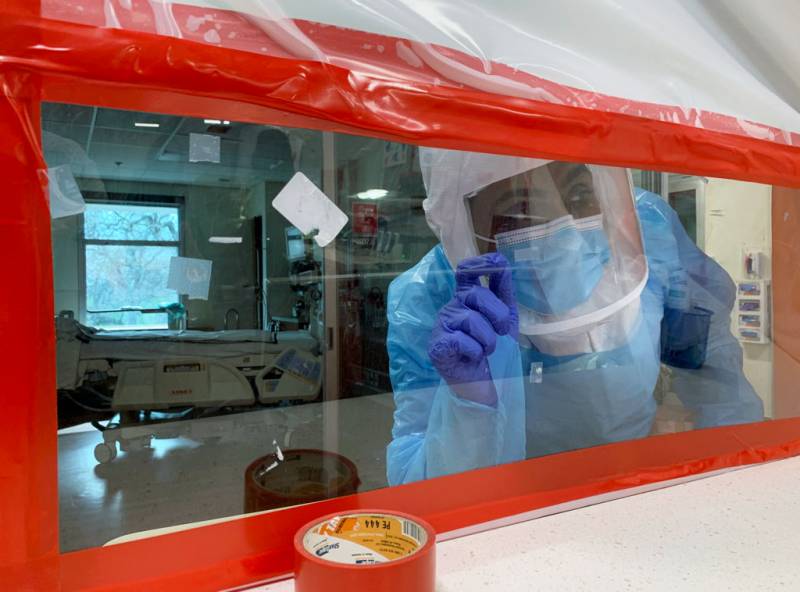 Someone in full personal protective equipment, including gloves and a hood with a clear face shield, looks through a plastic sheet taped to an opening in the wall with red duct tape.