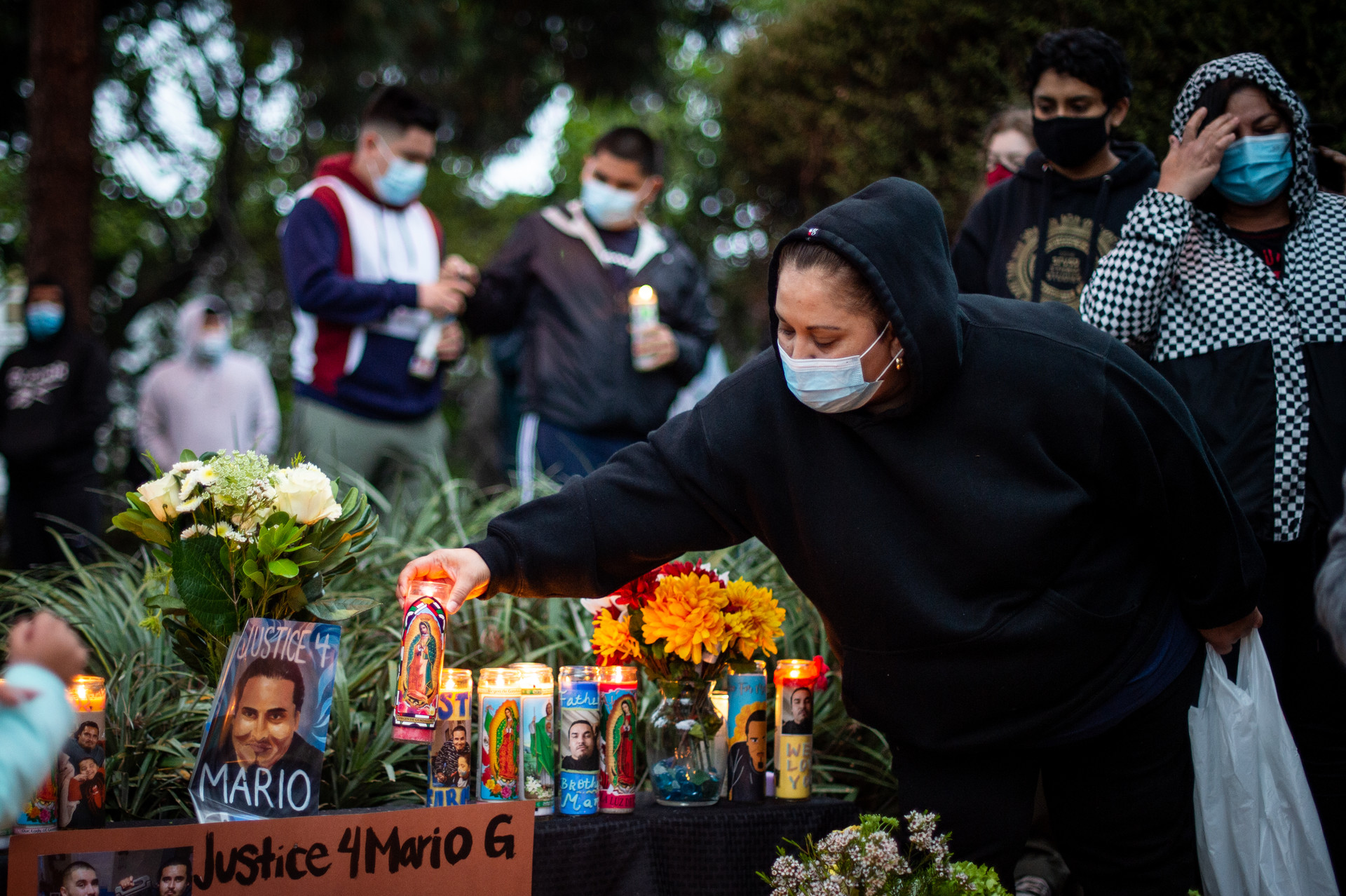 A woman wearing a black sweatshirt and a mask places a candle at an altar above the words "Justice 4 Mario G."