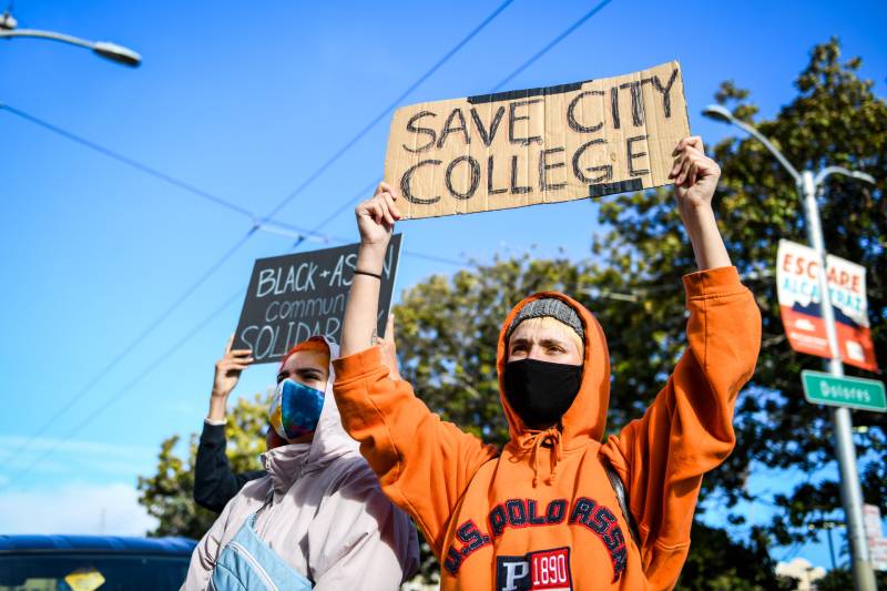 A person in an orange sweatshirt holds wearing a mask holds up a cardboard sign that says "Save City College" on a sunny day.