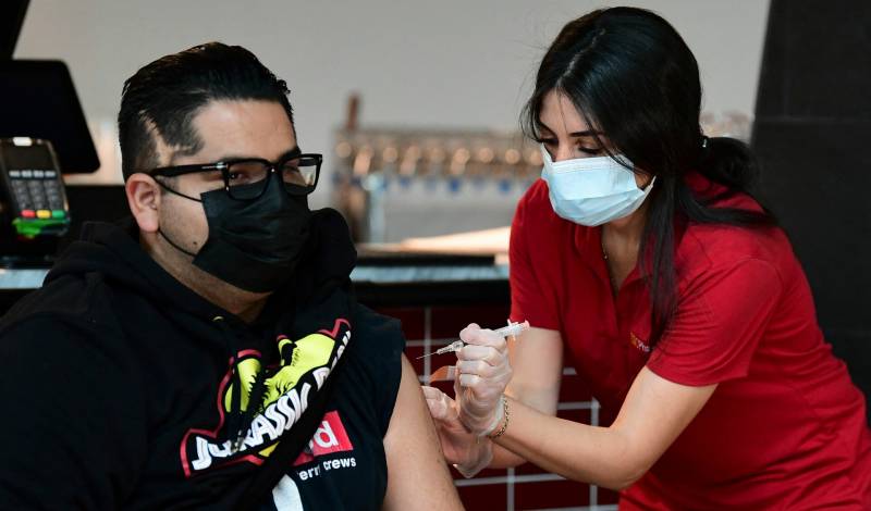 A young man wearing a Jurassic Park hoodie and glasses receives his vaccine from a pharmacist wearing a red shirt.