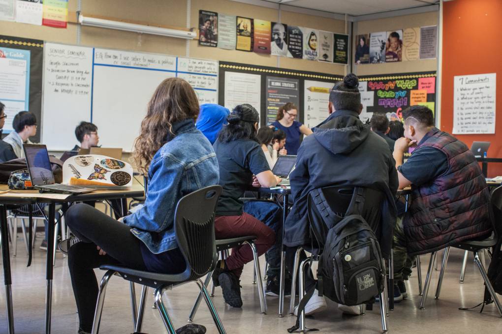 High school students sit on chairs behind tables facing a teacher, who stands.