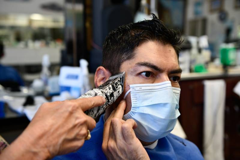 Carlos Hernández sostiene su cubrebocas mientras Sarah Cordiey le corta el pelo en Spiros Barber Shop en Long Beach, el 9 de junio de 2021.