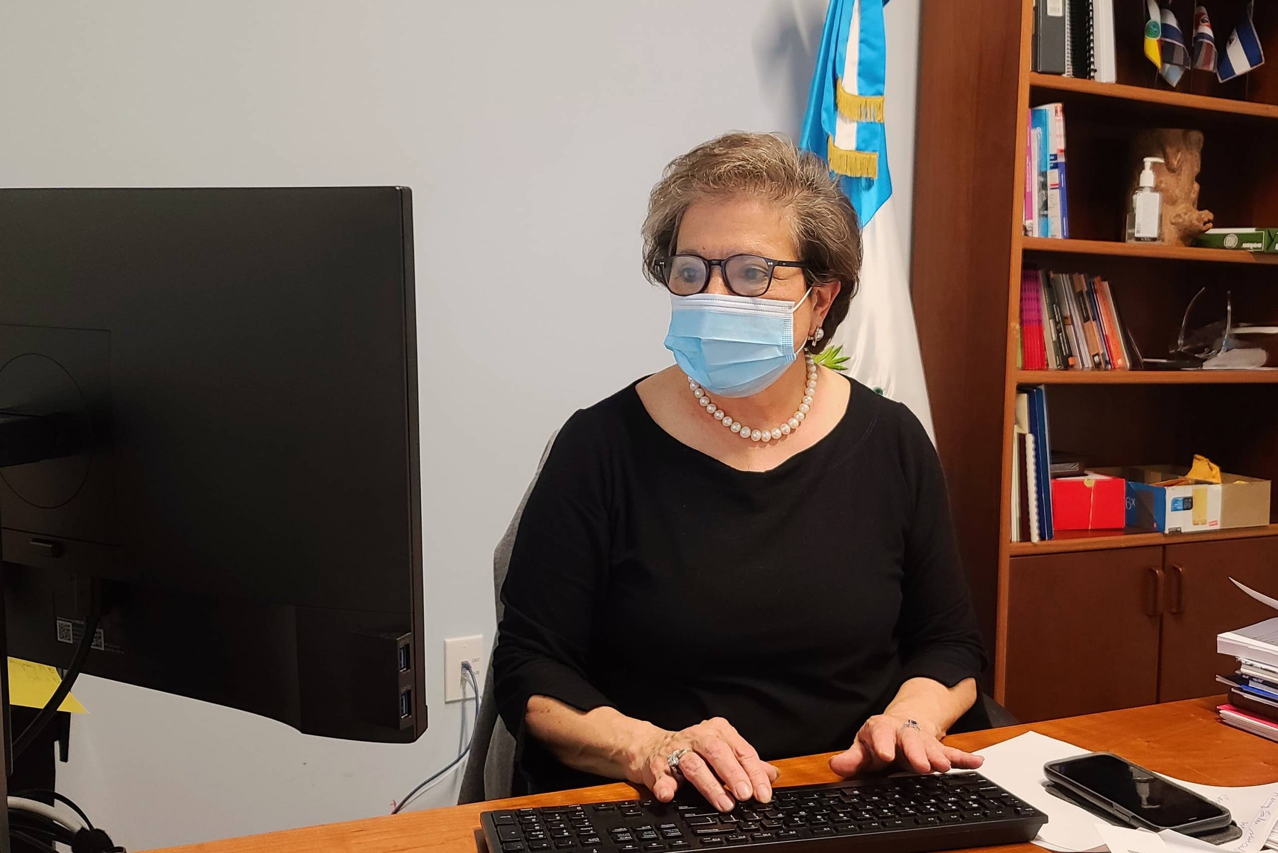 Sylvia Wohlers Gomar de Meie, the Guatemalan consul general in San Francisco, types on her computer in her office. She is wearing glasses and a blue facemask.