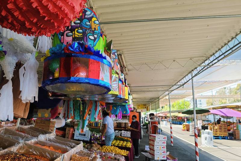 A piñata stand, with piñatas all around, next to a fruit and vegetable stand, some workers are visible moving boxes in the background.
