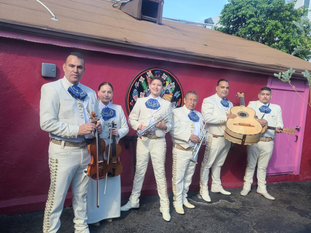 A group of mariachis stand against a wall with their instruments in their hands.