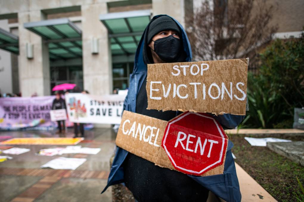 A person wearing a black face mask and a jacket stands outside holding signs that read "Stop Evictions" and "Cancel Rent."