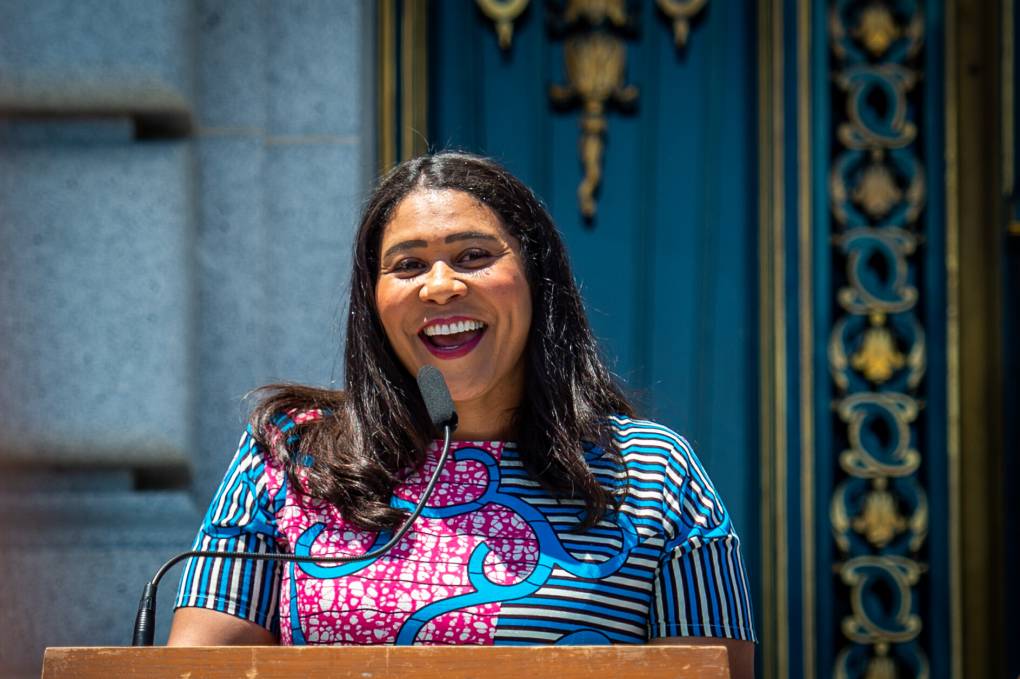 San Francisco Mayor London Breed speaks at a podium and smiles at a crowd.