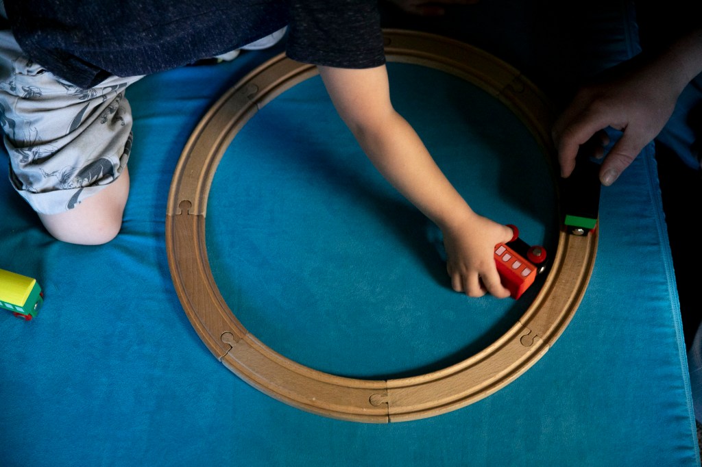 The camera views at the arm of a child who plays with a toy train. The toy train is red and made of wood and moves around wooden railroad tracks that are set in a circle.
