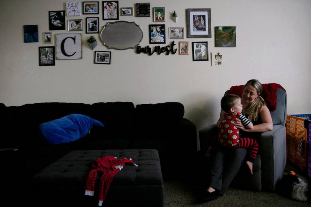 An adult with blond hair sits on a couch and carries an infant. Both are in a dark living room. There are many photos hung on the wall besides them.