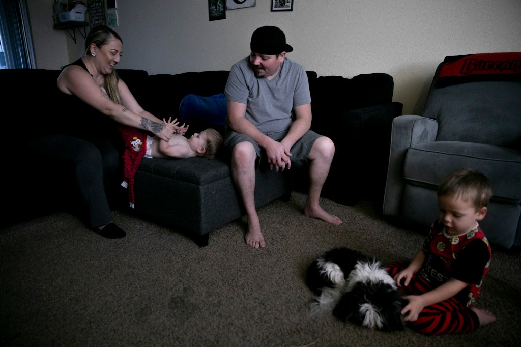 Two adults are sitting on the couch, looking at a baby, who is resting on the lap of one of the adults. Another young child plays with a dog on the carpet not too far from them.
